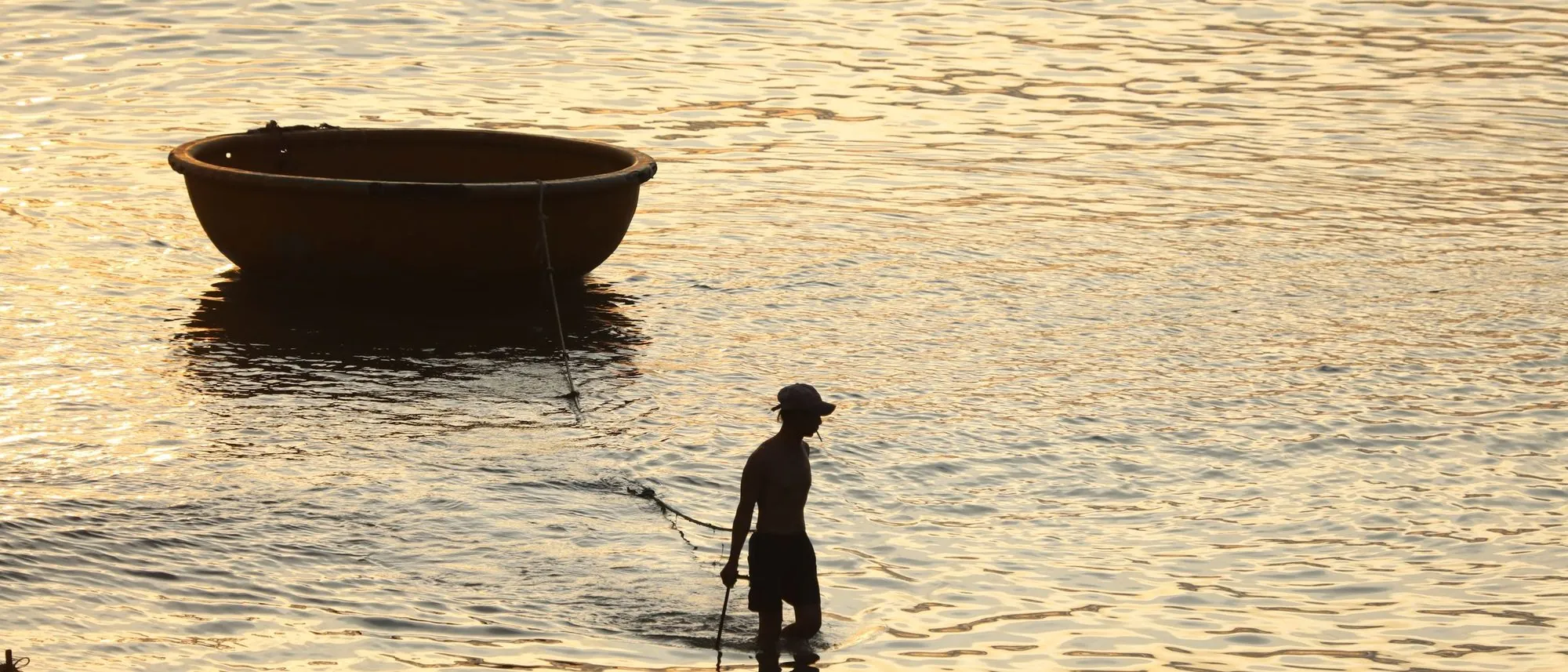 The silhouette of a fisherman wading through water at sunset pulling a round coracle behind him.