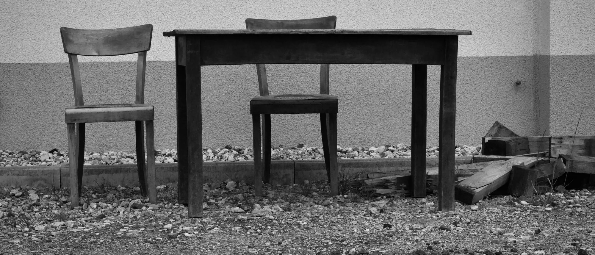 Two worn wooden chairs and a small table stand against a wall outdoors in black and white.