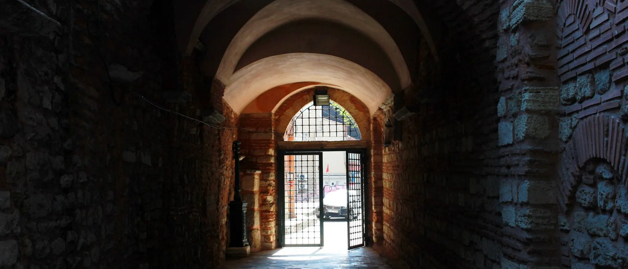 A dim stone corridor of arches leading towards a gated opening and daylight.