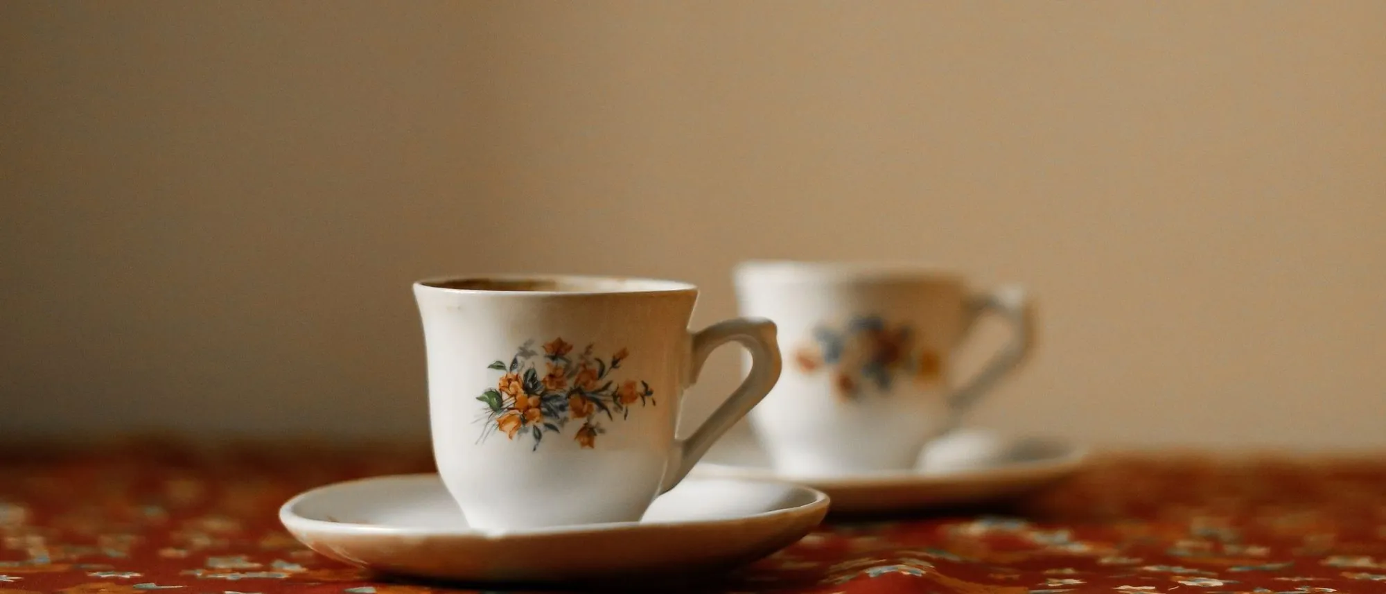 Two floral teacups sit on a richly patterned tablecloth.