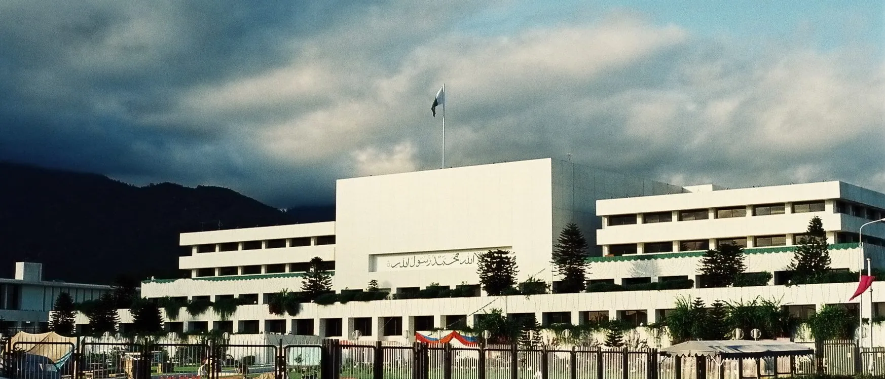 Parliament House, Islamabad, a white modernist block under a stormy sky with the Margalla Hills behind