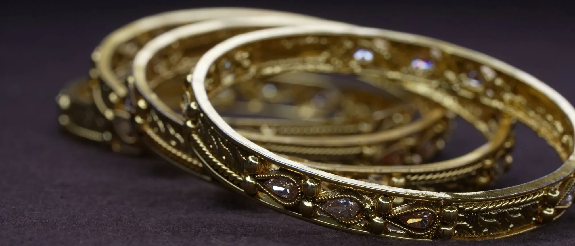 Three gold bangles with fine filigree and small stones resting on a dark cloth