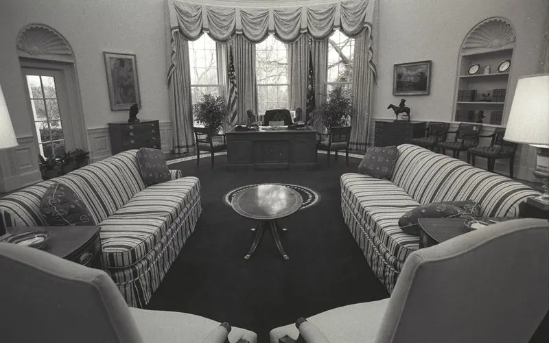 The Oval Office empty, photographed in black and white: the desk before the windows, two striped sofas facing each other and no one in the room