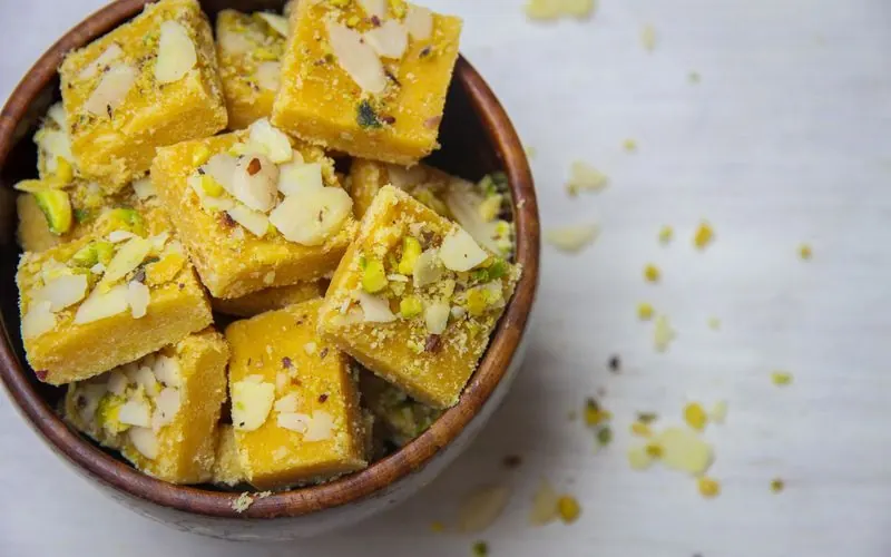 Pieces of besan barfi topped with almonds and pistachios in a wooden bowl seen from above.