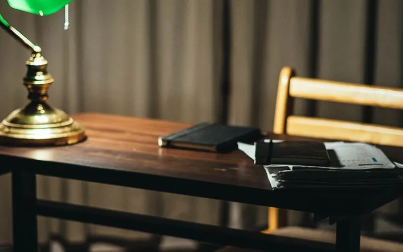 A dark room with a desk, an empty chair and one lit green banker's lamp
