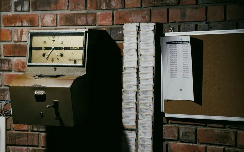 A retro punch-clock and rack of time cards on a dark brick wall