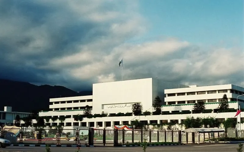 Parliament House, Islamabad, a white modernist block under a stormy sky with the Margalla Hills behind