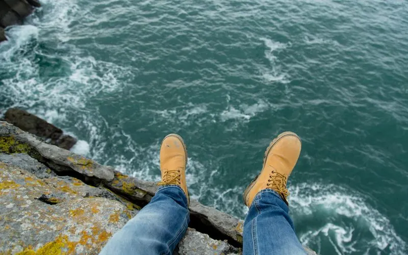 A person in boots sits at the edge of a sea cliff looking out over the ocean.