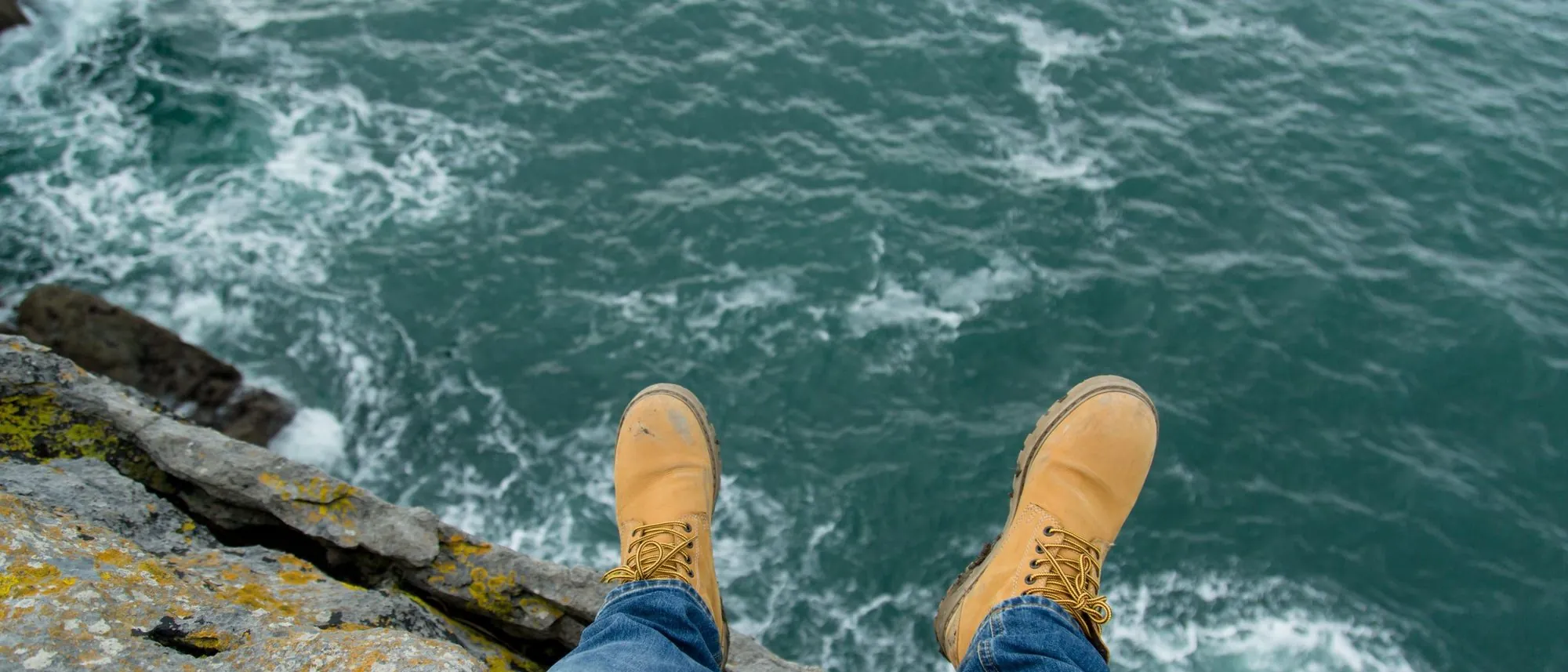 A person in boots sits at the edge of a sea cliff looking out over the ocean.