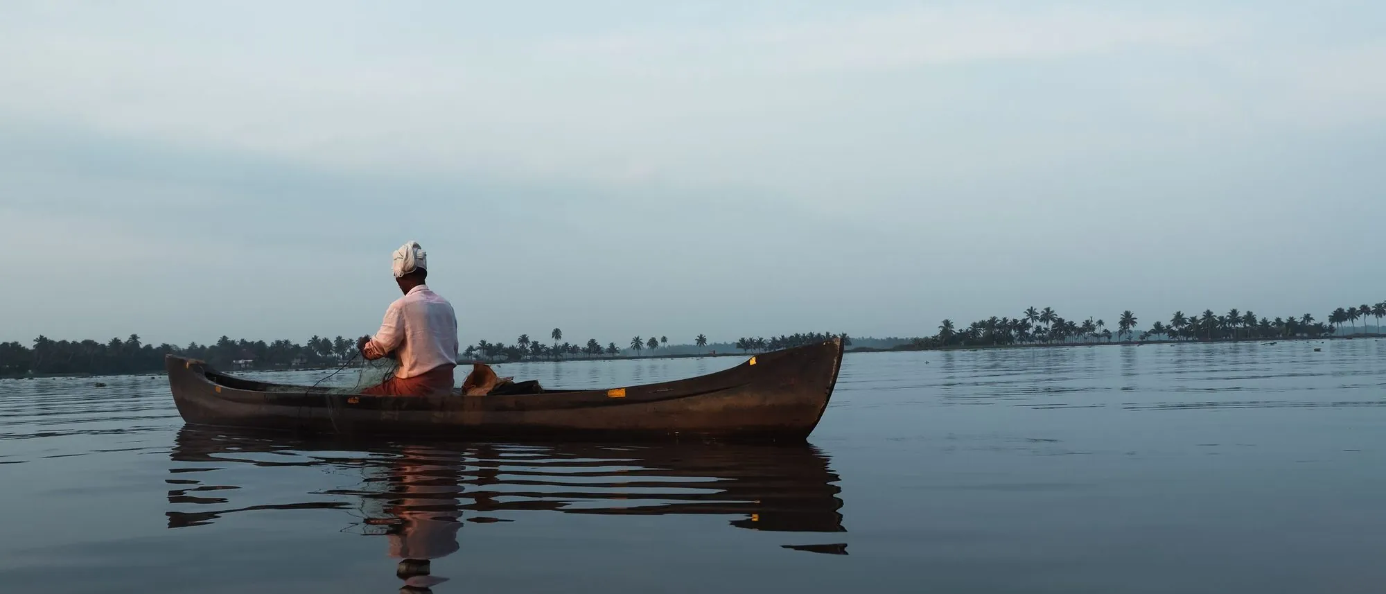 A man paddles a canoe across the still Alappuzha backwaters at dawn.