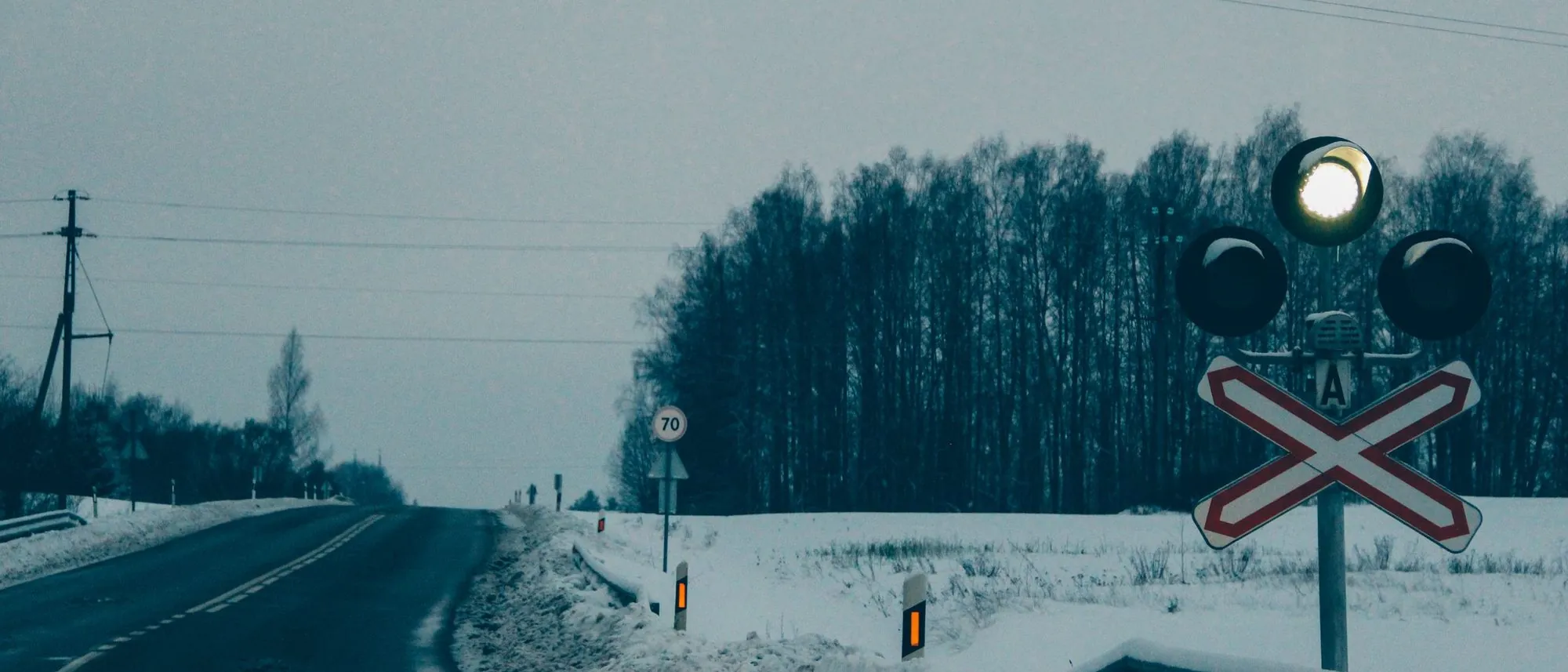 A railway crossing signal beside a snowy rural road with forest behind.