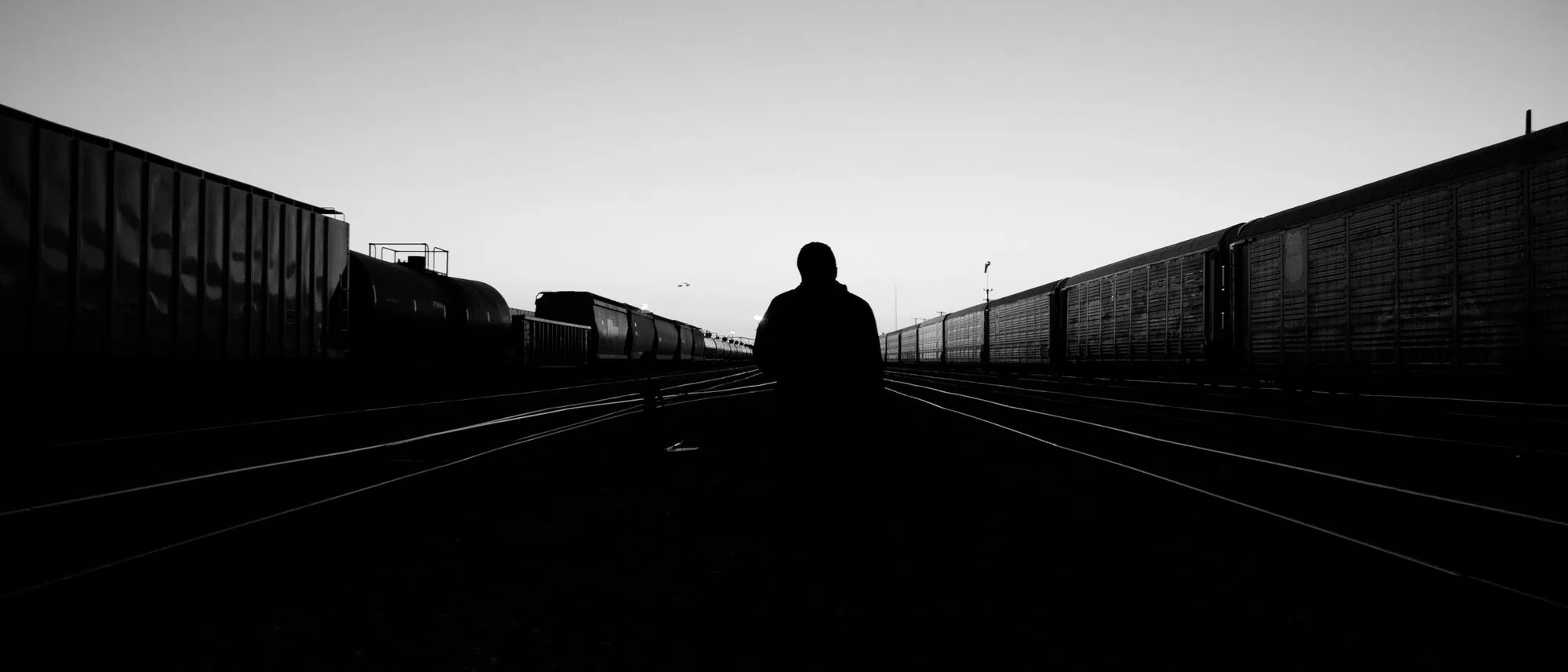 A lone silhouetted figure walks along an empty railway track at dusk in black and white.