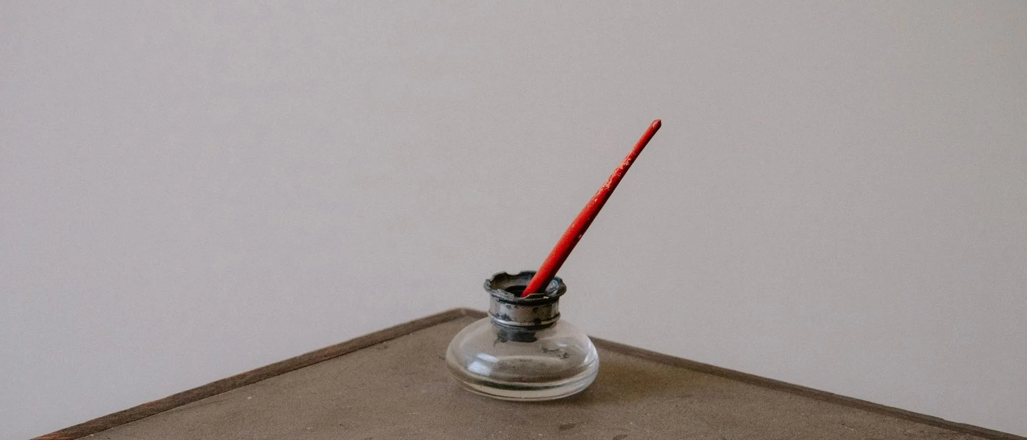 A glass inkwell with a red pen resting in it on a rustic wooden desk.